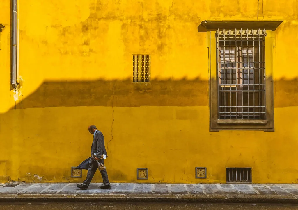 A man walking down a street passing in front of a bright yellow building